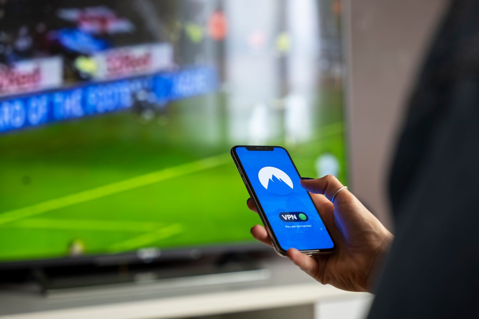 Hand hält ein Smartphone mit verbundener VPN-App vor einem Fernseher, auf dem ein Fußballspiel läuft