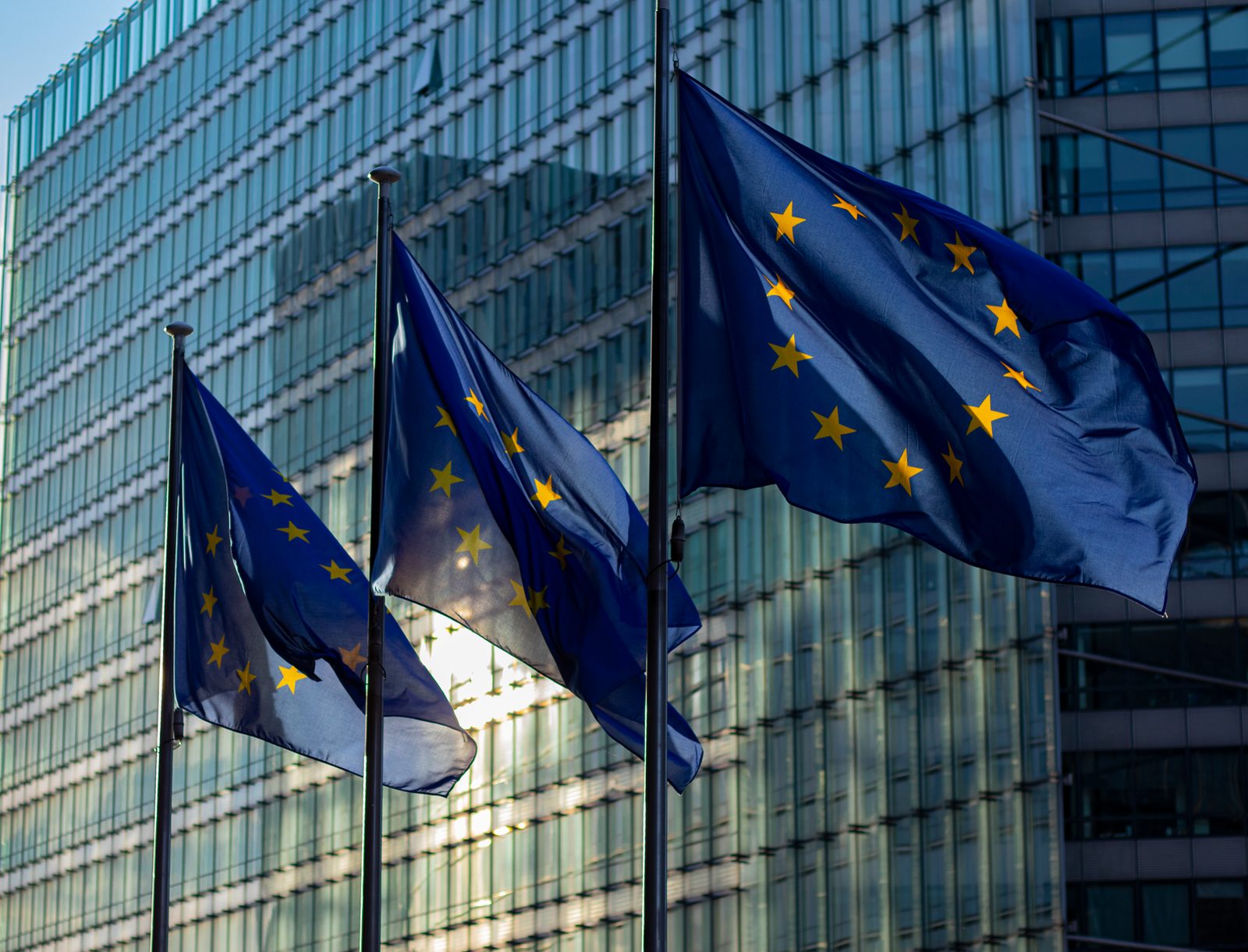 Tre bandiere europee che sventolano davanti alla facciata vetrata di un edificio istituzionale a Bruxelles