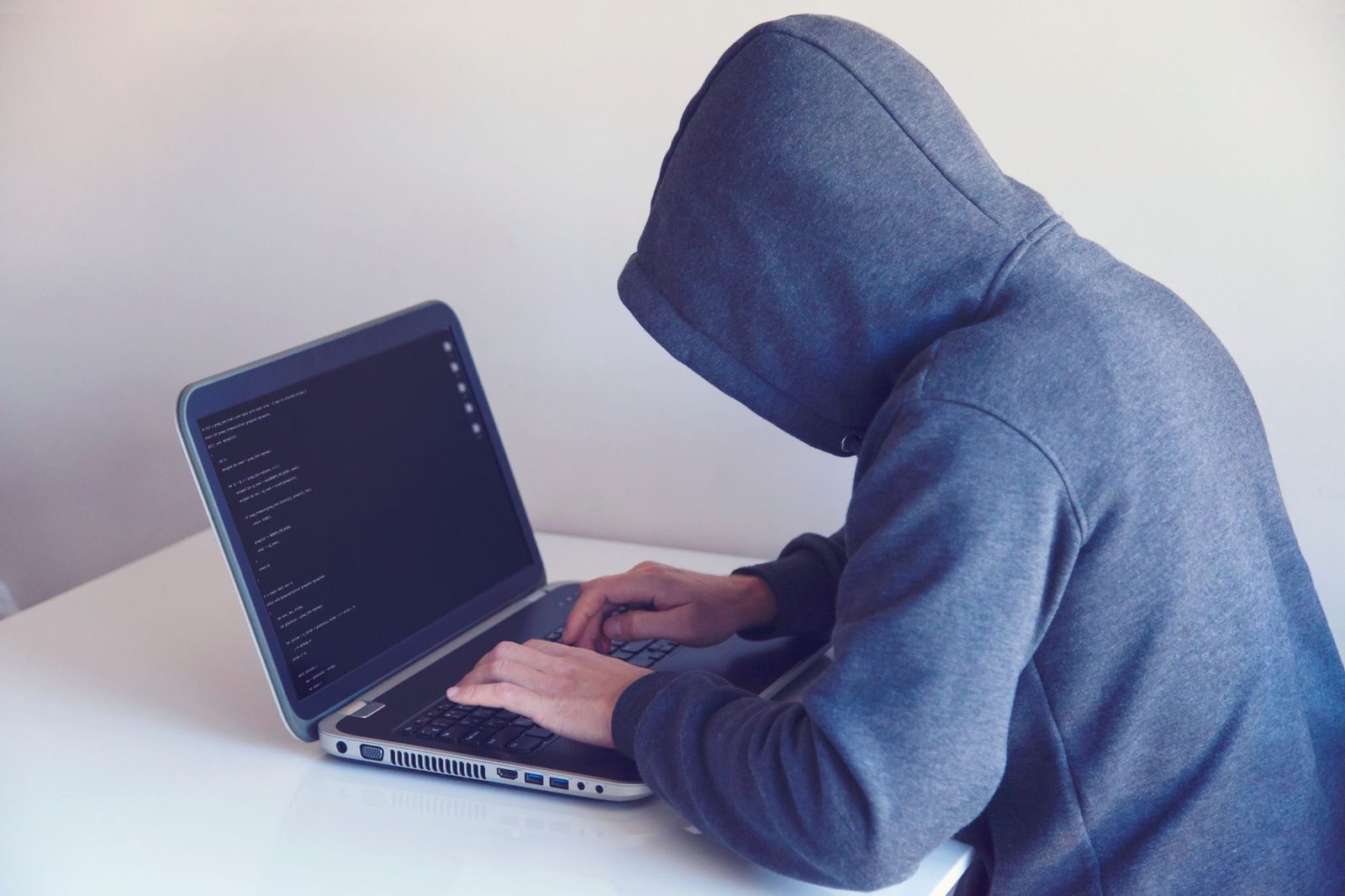 Personne au visage caché sous une capuche grise tapant du code sur un ordinateur portable, à un bureau blanc