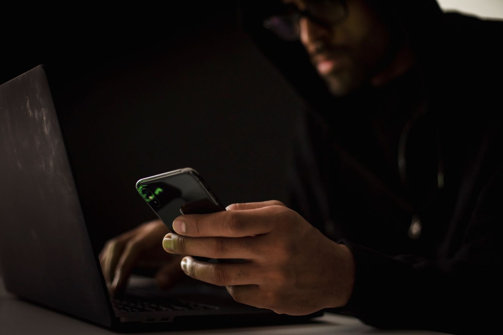 Homme à capuche dans la pénombre utilisant un smartphone devant un ordinateur portable