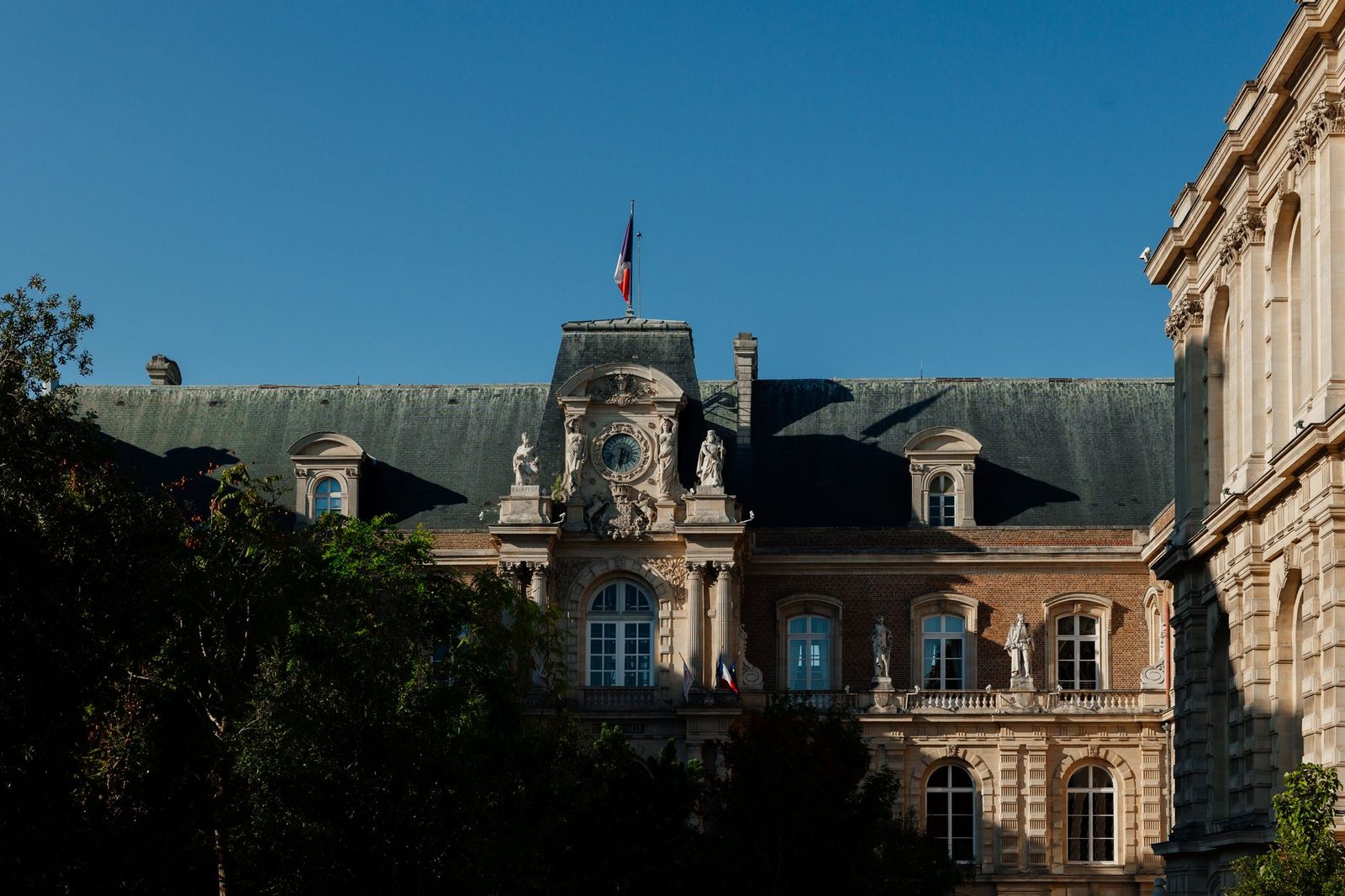 Façade d'un bâtiment administratif français en pierre et brique surmonté d'un drapeau tricolore, ciel bleu