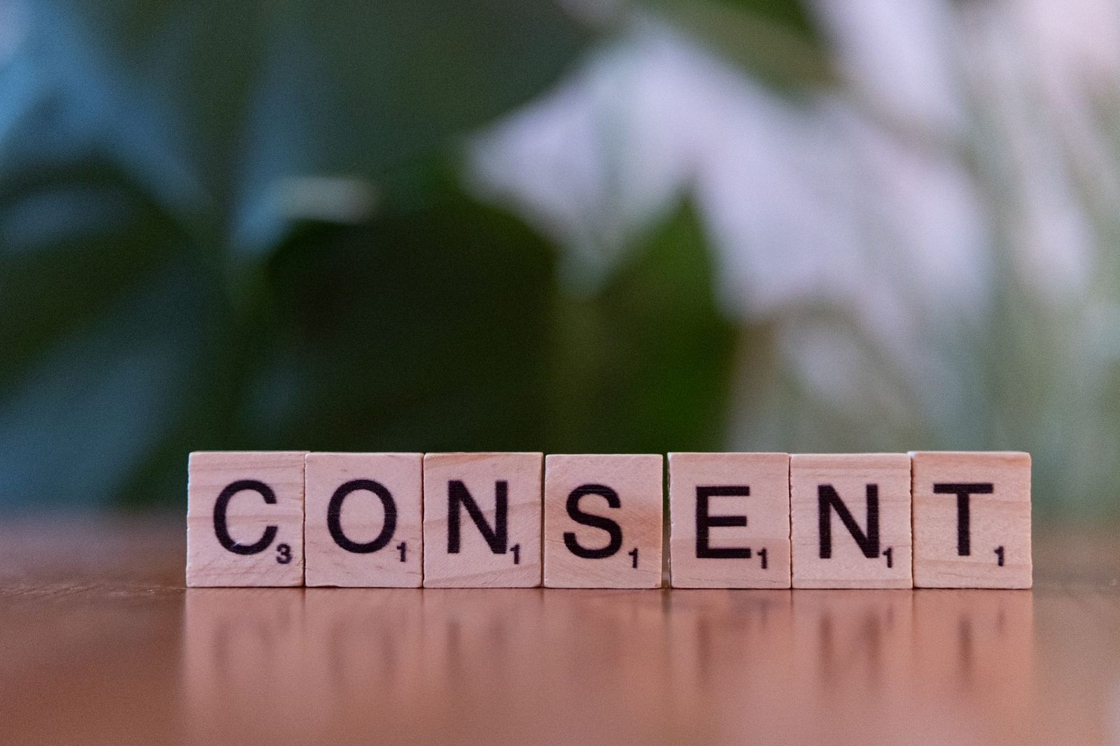 Lettres de Scrabble en bois formant le mot « CONSENT » sur une table, feuillage vert flou en fond