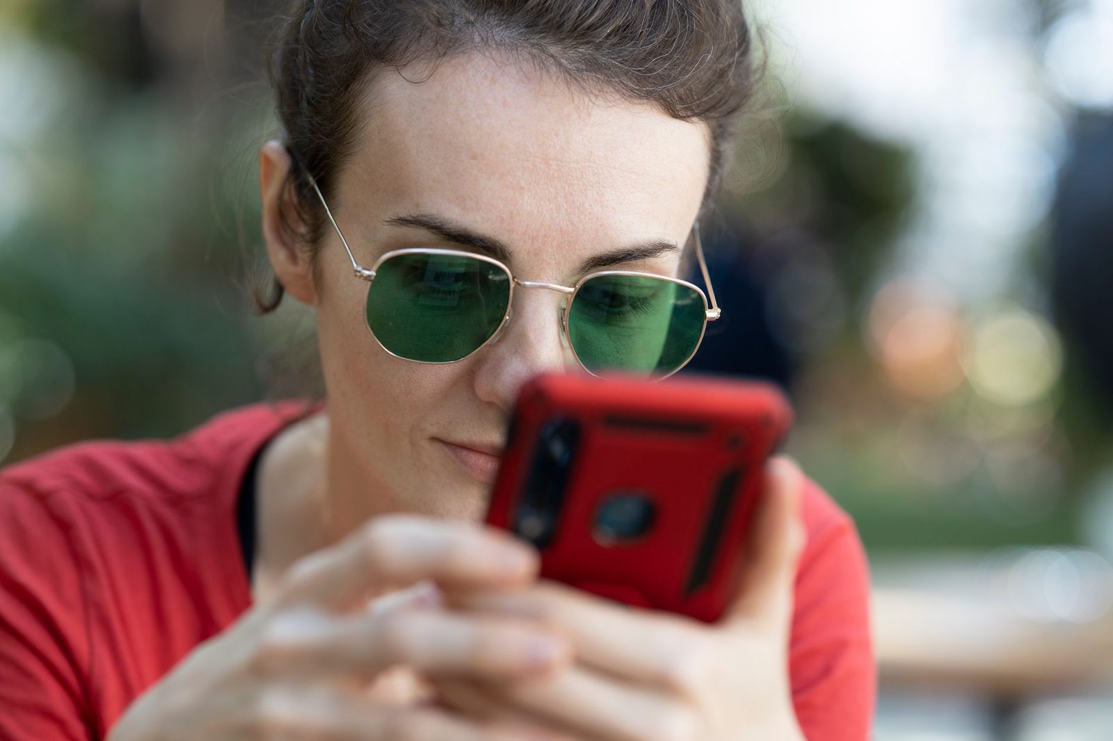 Woman wearing green sunglasses looking closely at her smartphone in a red case