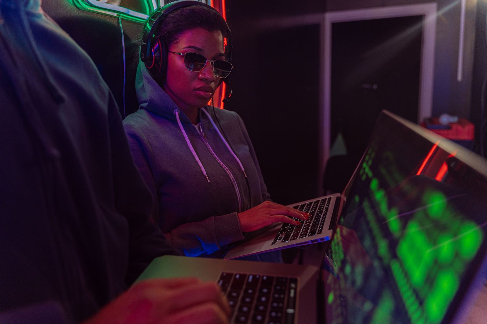 Woman in a hoodie and glasses typing on a laptop in a dark room lit by neon lights