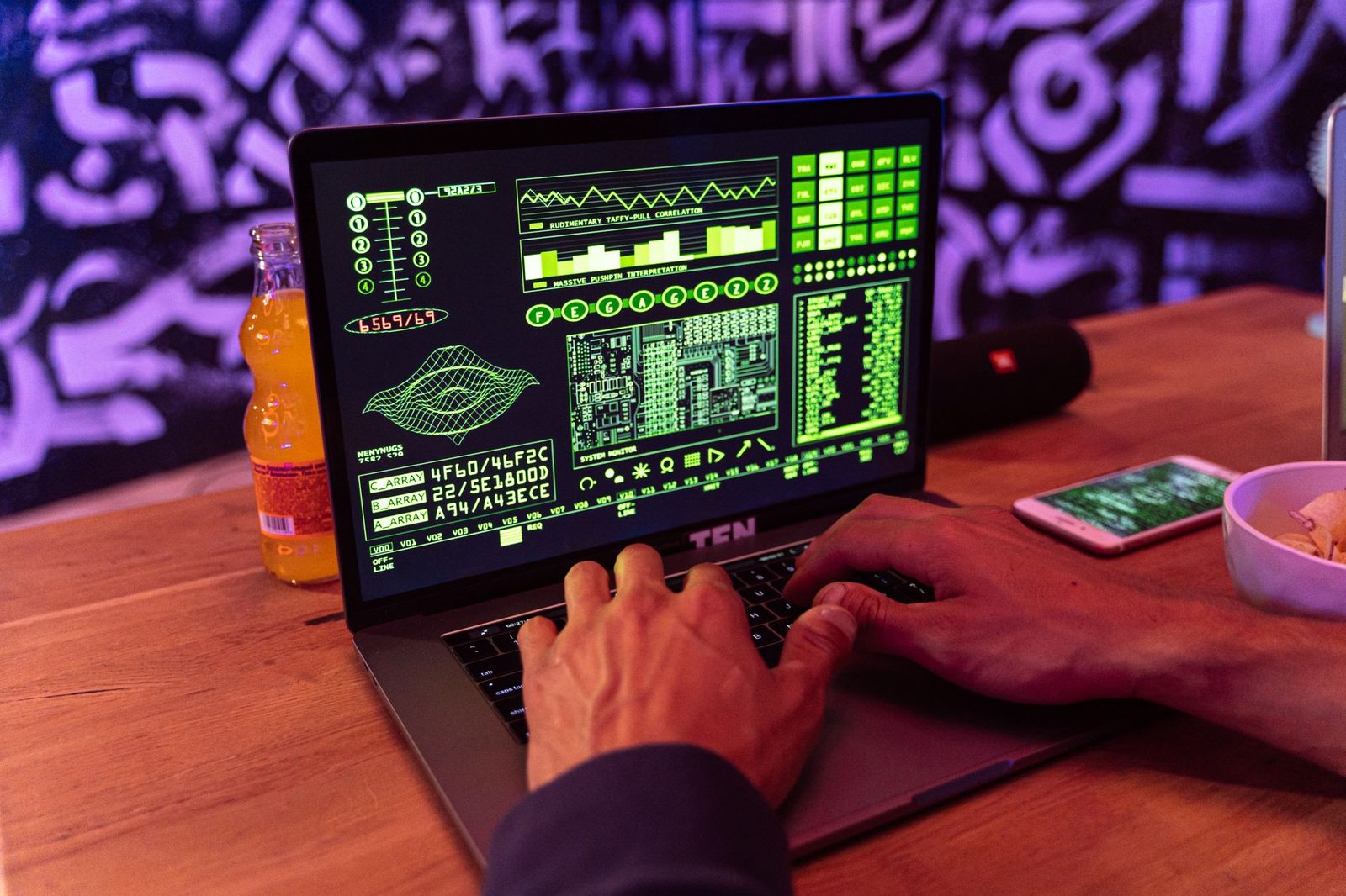 Mani che digitano su un computer portatile con dati verdi sullo schermo, smartphone appoggiato accanto su un tavolo