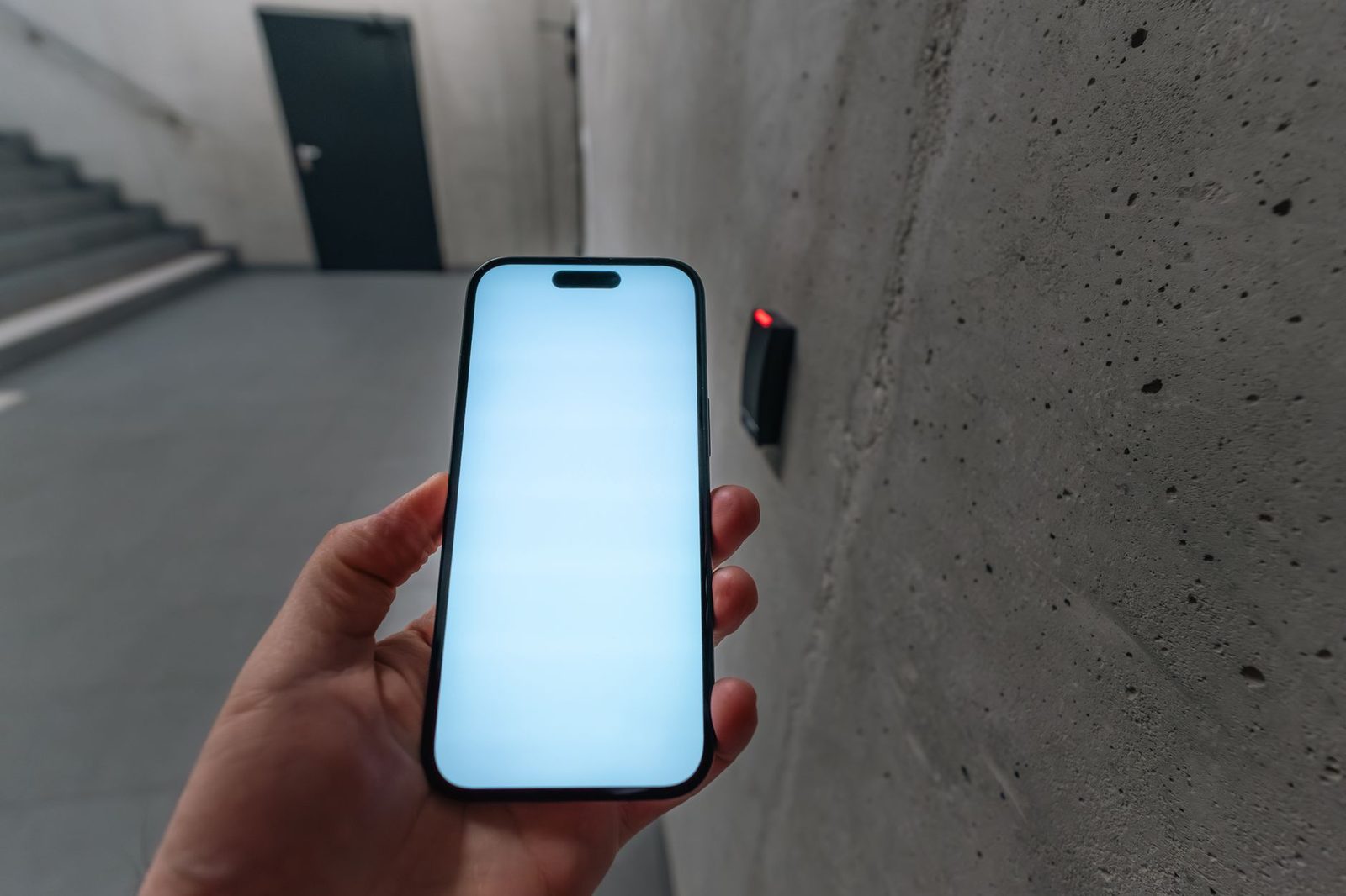 Mano che tiene uno smartphone con schermo vuoto davanti a un lettore di badge con LED rosso su un muro di cemento