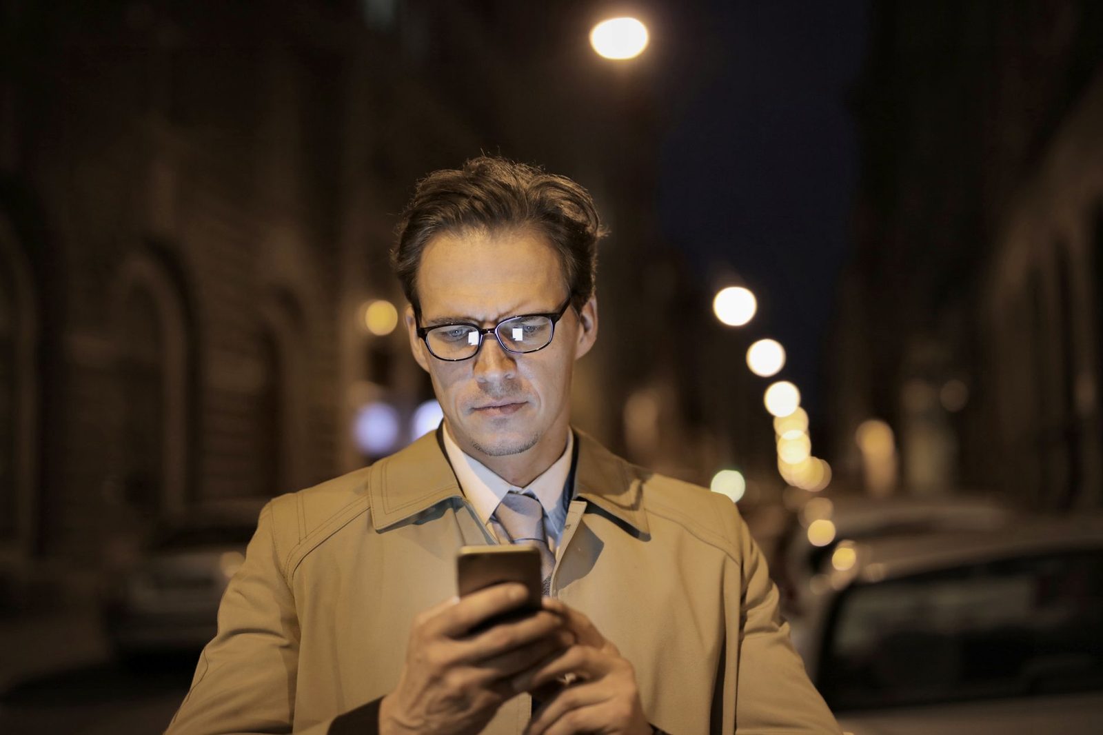 Homme à lunettes en trench-coat consultant son smartphone dans une rue de ville éclairée la nuit