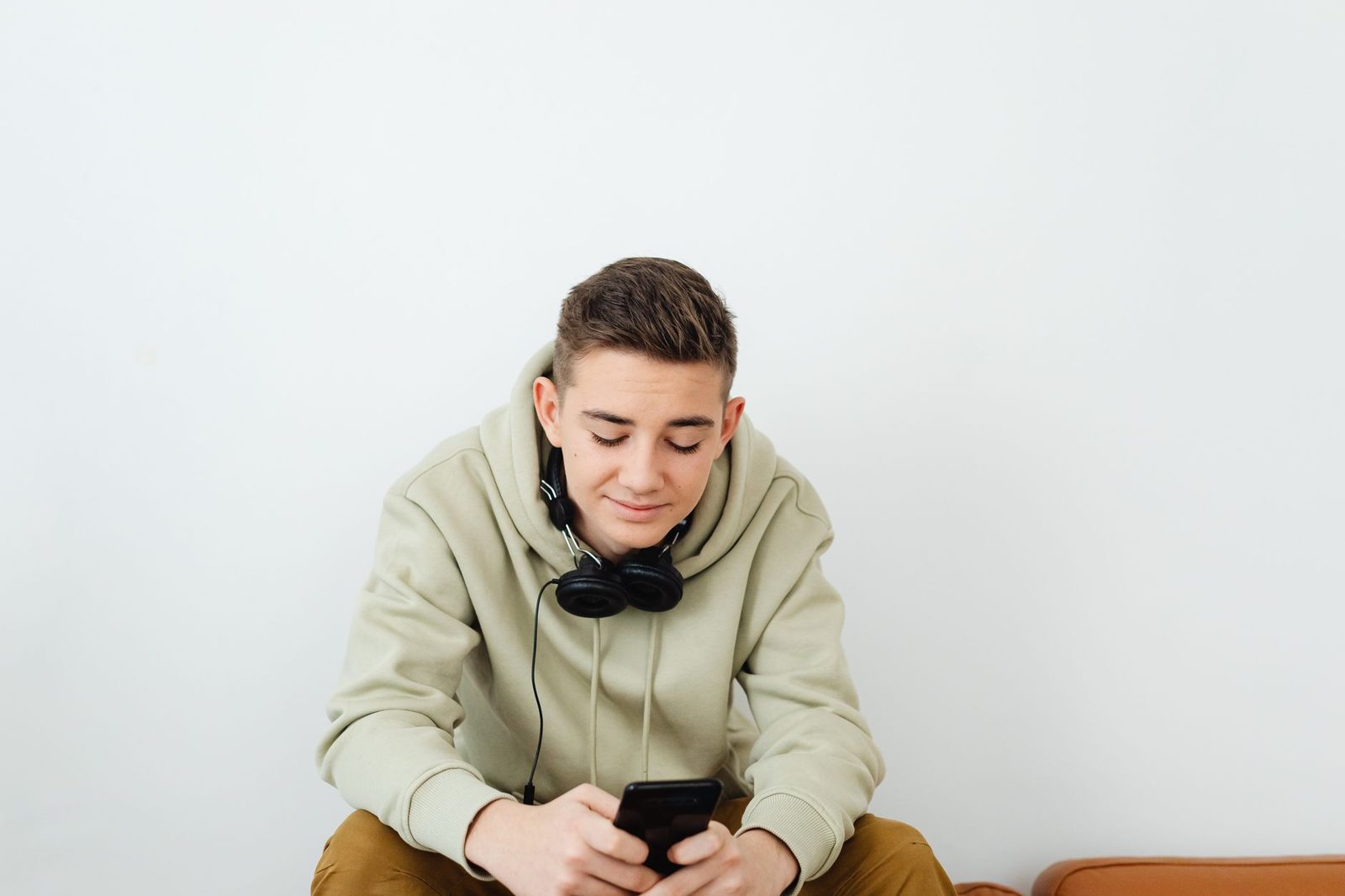 Adolescent en sweat à capuche, casque autour du cou, consultant son smartphone assis devant un mur blanc