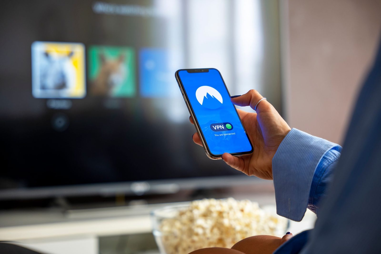 Hand holding a smartphone displaying a connected VPN app, in front of a television and a bowl of popcorn