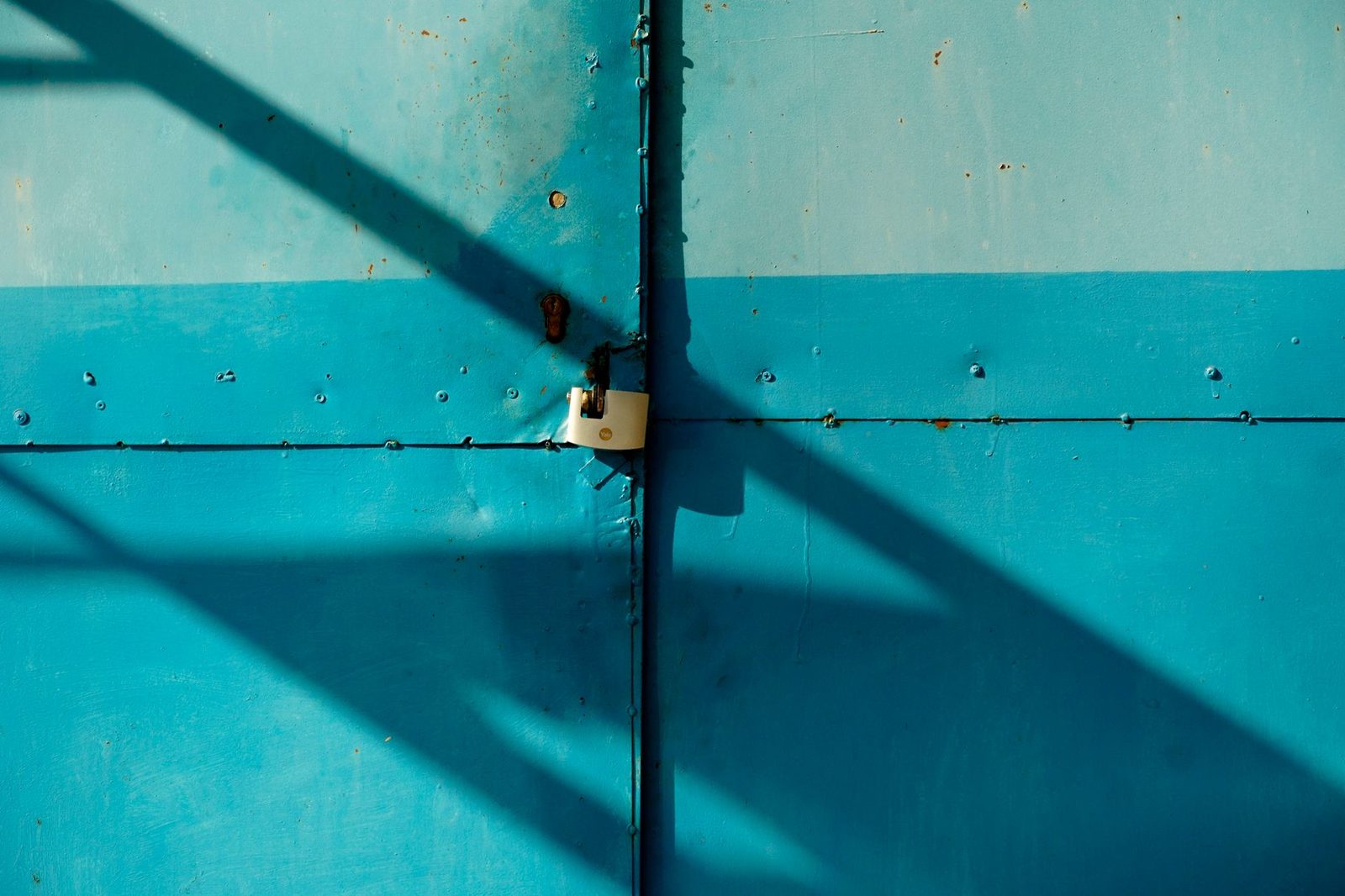 White padlock closed on a turquoise blue metal door crossed by diagonal shadows
