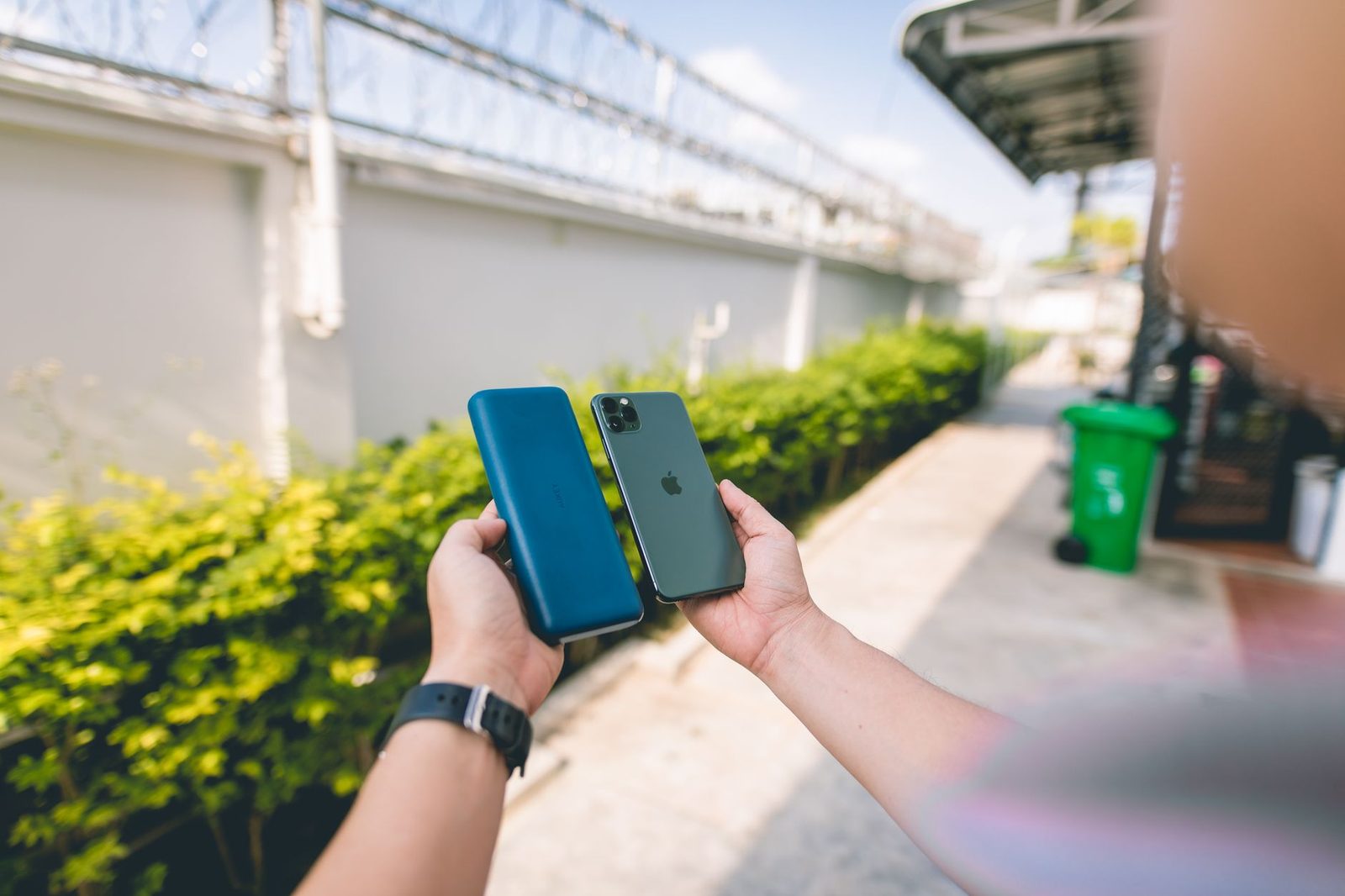 Main tenant un iPhone vert et une batterie externe bleue en extérieur, sur un trottoir bordé de haies