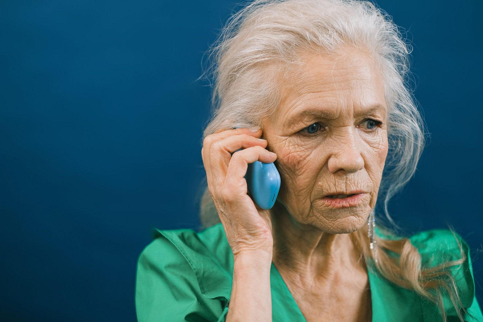 Elderly white-haired woman looking annoyed, blue phone to her ear, against a dark blue background