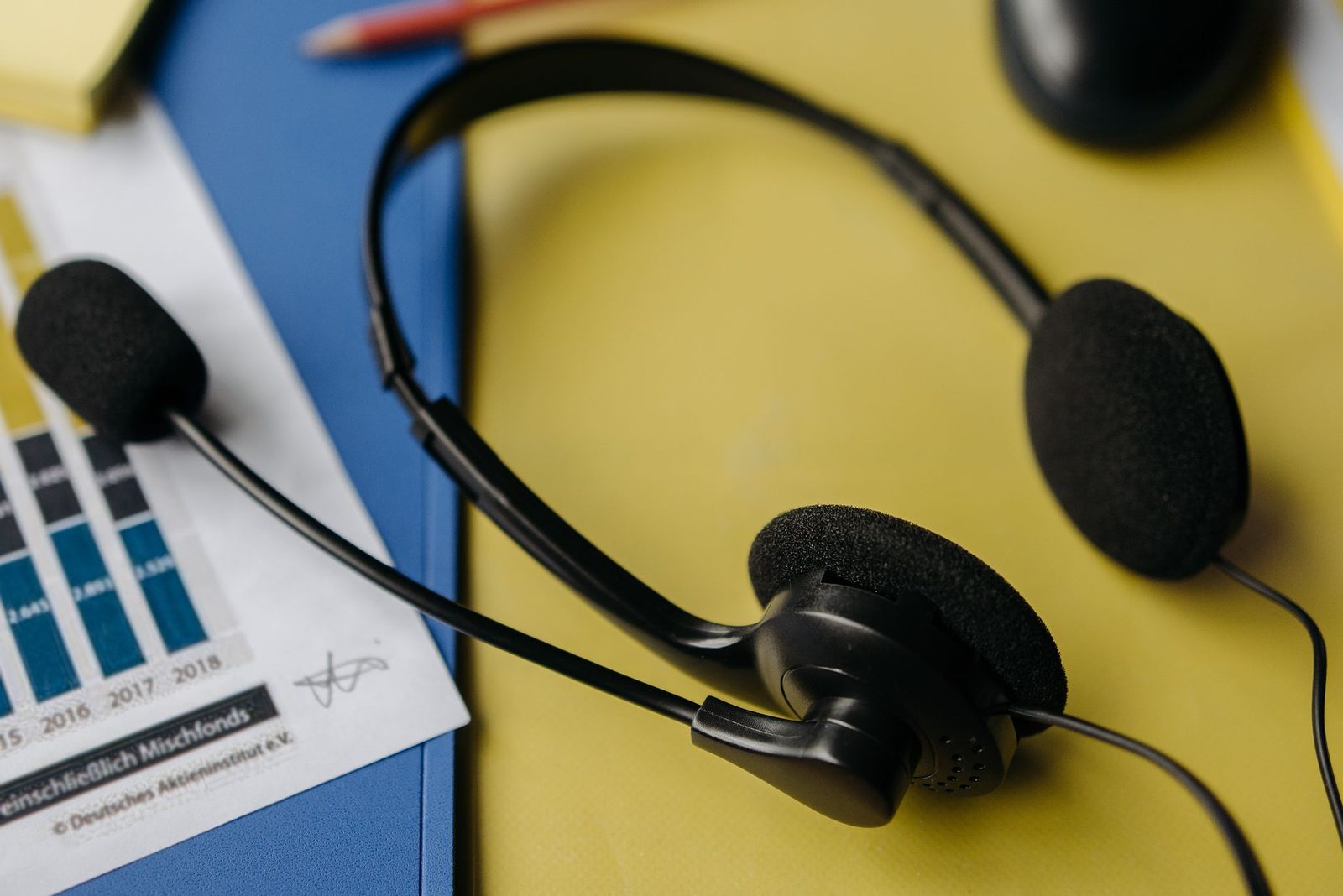 Black telephone headset resting on a yellow desk, next to a printed document and a blue folder