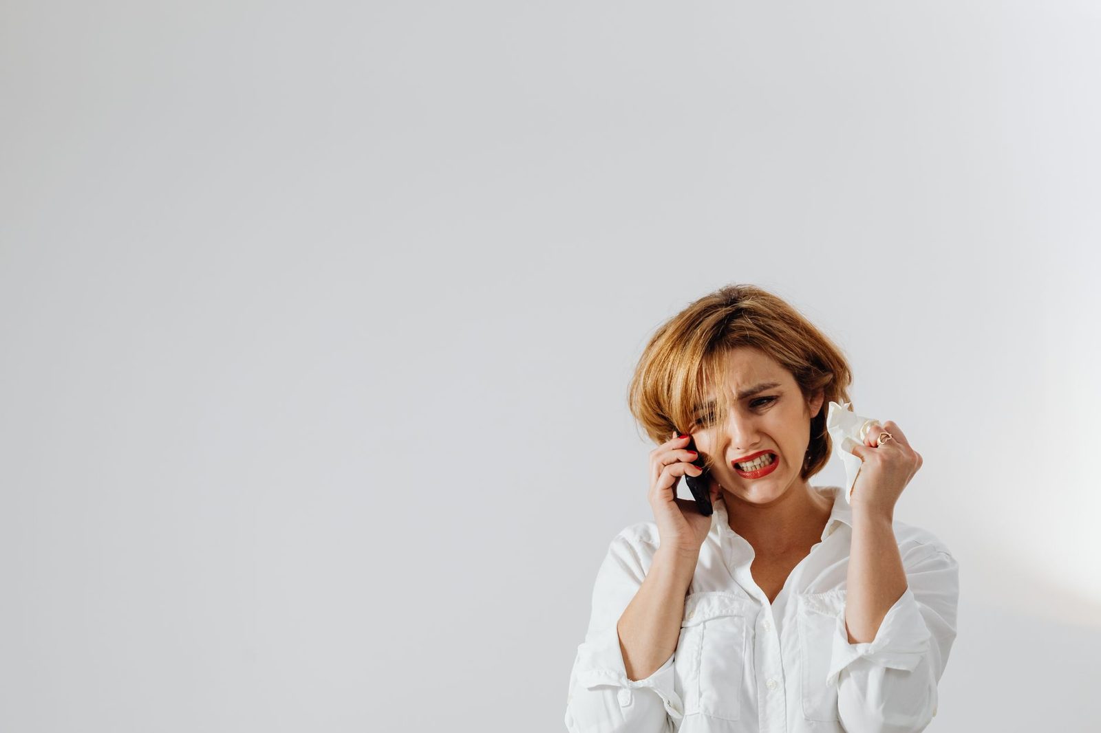 Young woman in a white shirt, looking irritated, on her mobile phone in front of a white wall