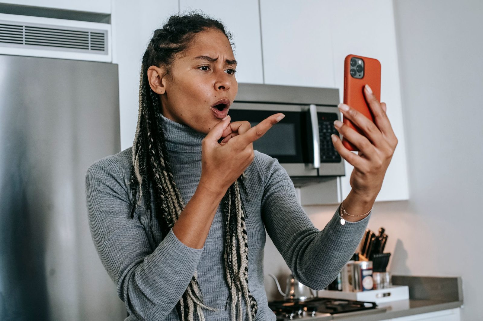 Annoyed woman in a kitchen pointing at the red smartphone she is holding up