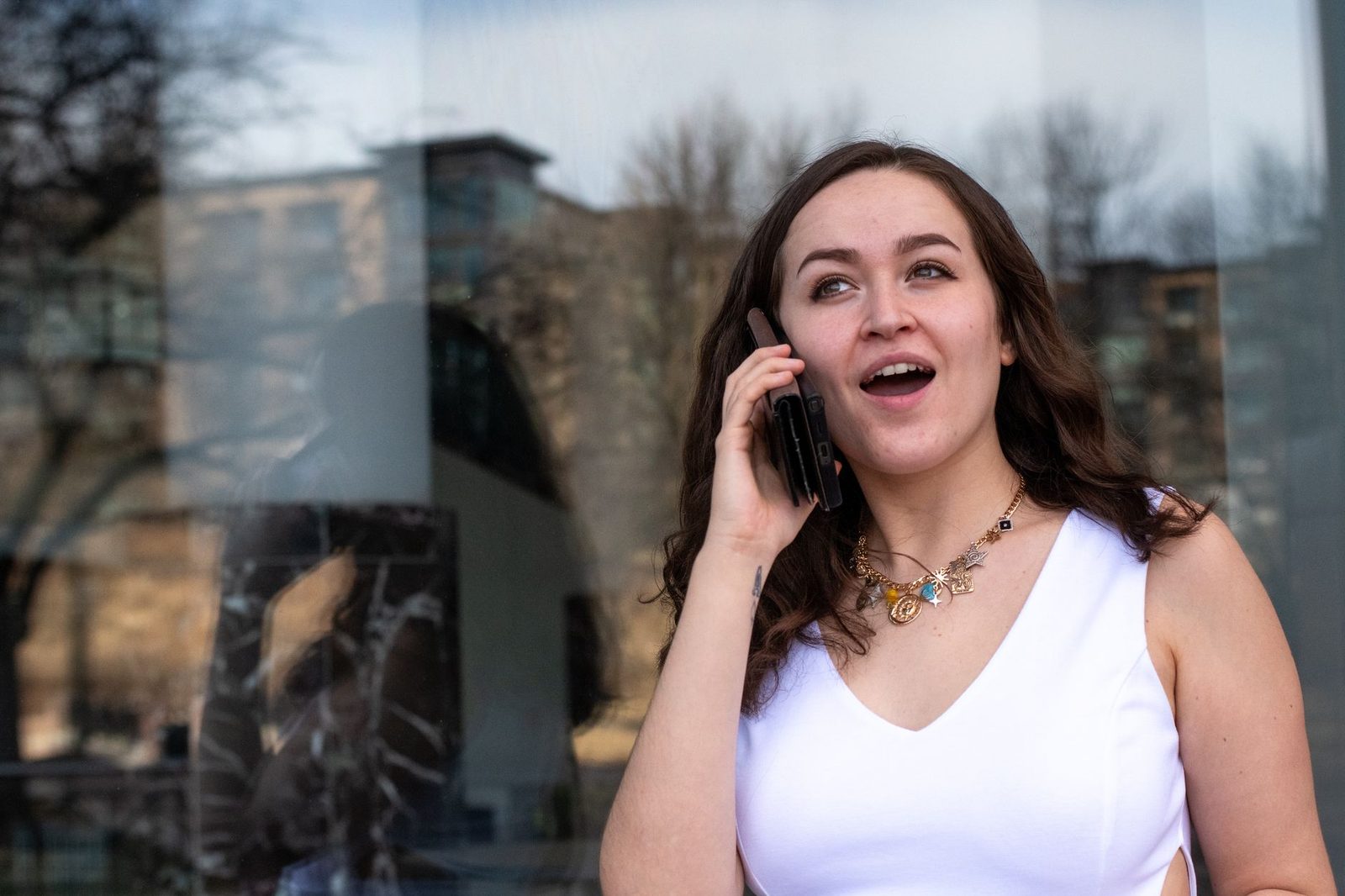 Smiling young woman in a white top talking on a smartphone in front of a glass façade