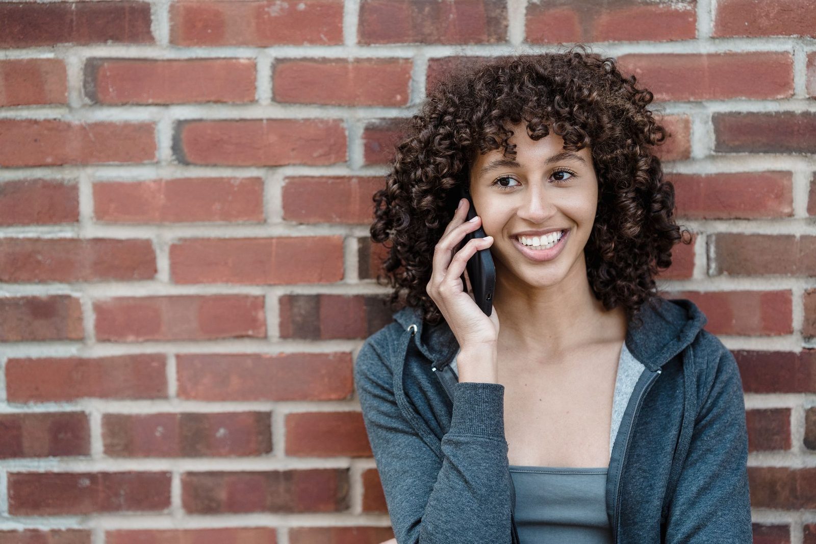 Smiling young woman with curly hair on a smartphone in front of a red brick wall