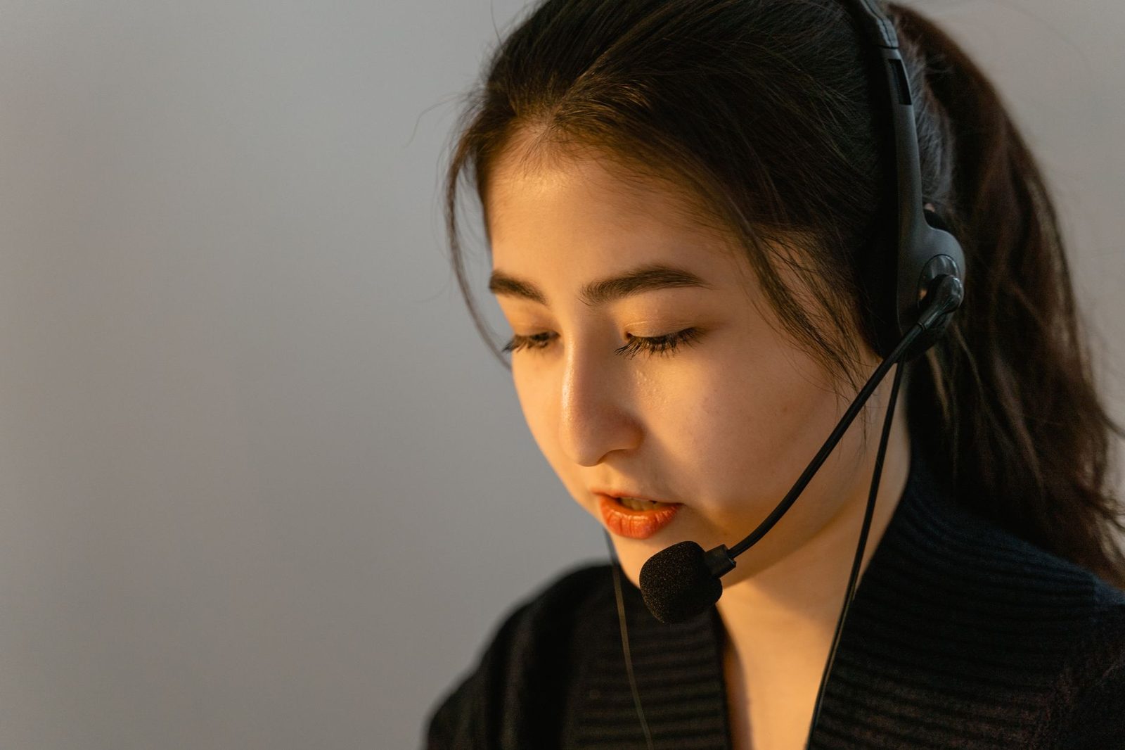 Young woman wearing a call-centre headset, eyes lowered, in front of a grey wall