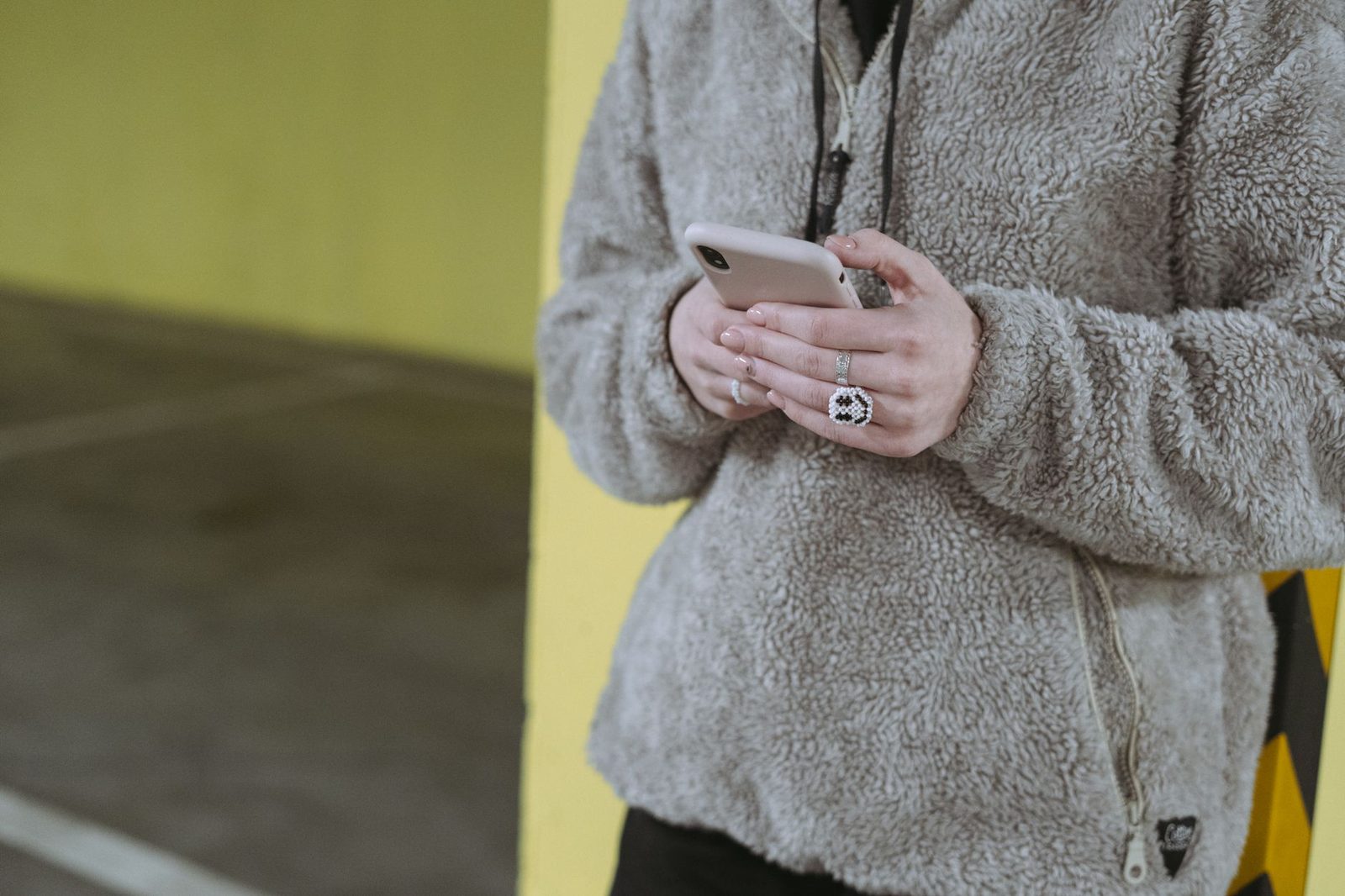 Femme en veste polaire grise tenant un smartphone dans les mains, adossée à un mur jaune