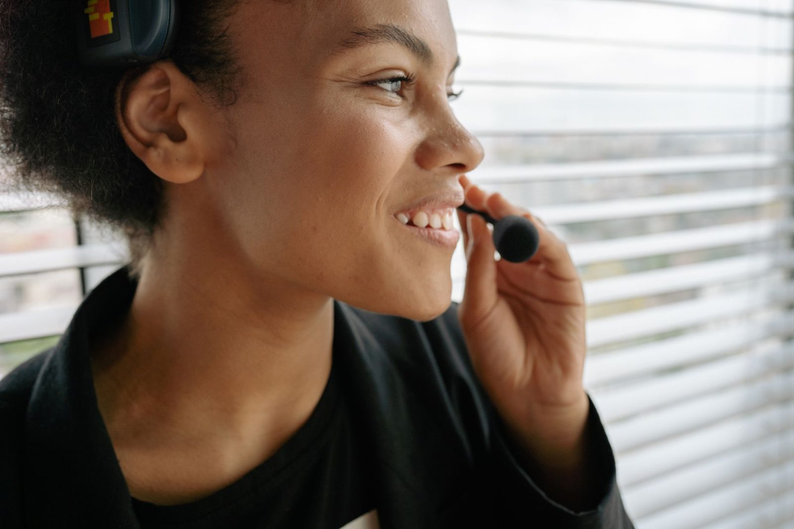 Smiling young woman wearing a call-operator headset by a window with blinds