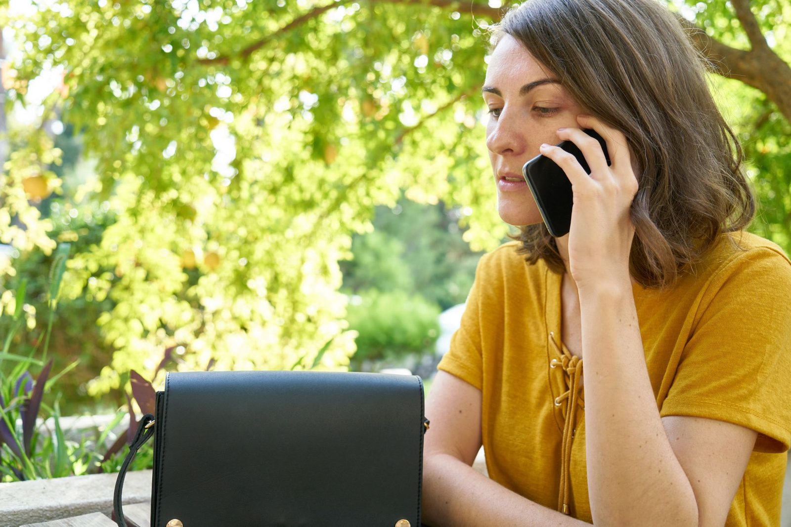 Woman in a yellow T-shirt on the phone outdoors, looking worried, black handbag in front of her