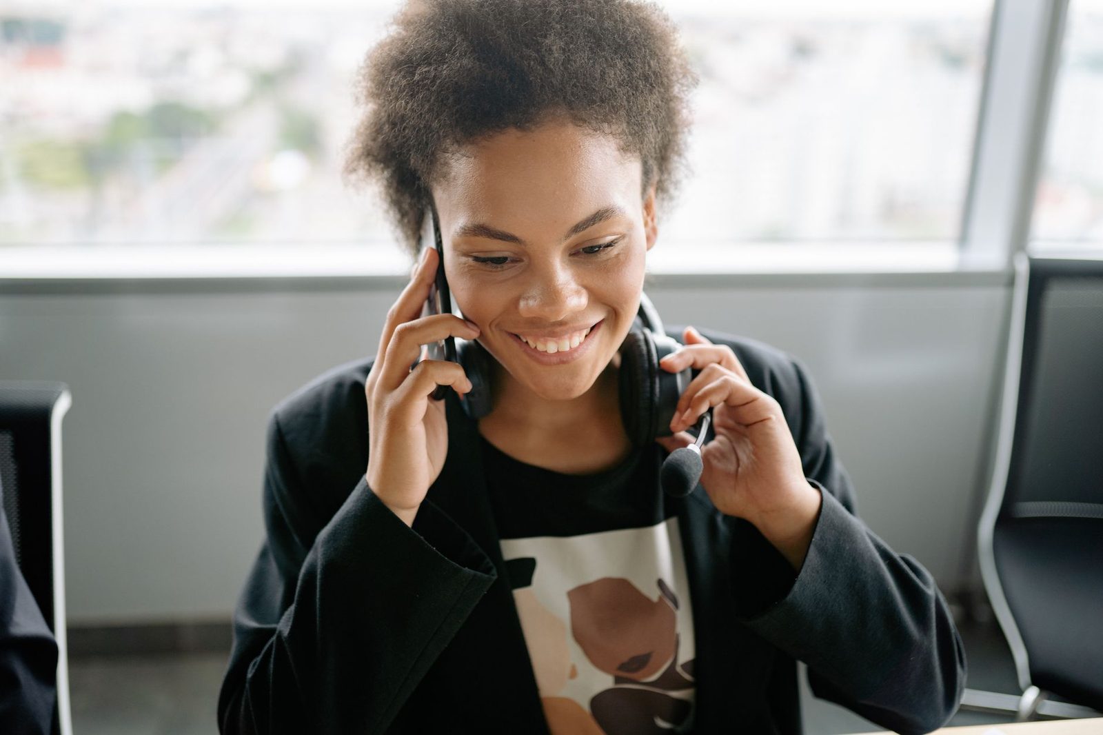Smiling young woman on a mobile phone, headset around her neck, sitting in a bright office